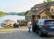 Woman with a tablet standing beside a blue SUV in a gravel driveway in front of a lakeside house with a dock and boat visible by the water.