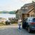 Woman with a tablet standing beside a blue SUV in a gravel driveway in front of a lakeside house with a dock and boat visible by the water.