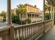 Wide porch of a historic beige house with white columns, palm trees, and a cobblestone street.
