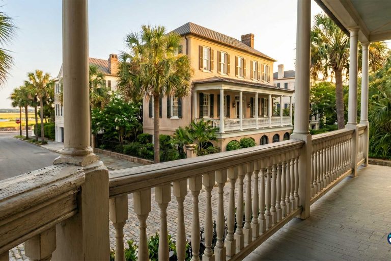 Wide porch of a historic beige house with white columns, palm trees, and a cobblestone street.