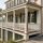 An elevated, luxury Charleston-style coastal home with immaculate cream-colored walls, dark green shutters, and crisp white porch railings. The property features a polished wooden deck, brand-new copper gutters, and a palm tree, all overlooking a serene salt marsh with winding waterways under a bright sky.