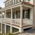 An elevated, luxury Charleston-style coastal home with immaculate cream-colored walls, dark green shutters, and crisp white porch railings. The property features a polished wooden deck, brand-new copper gutters, and a palm tree, all overlooking a serene salt marsh with winding waterways under a bright sky.