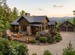 An aerial, wide-angle view of a luxury rustic mountain home with dark wood siding, stone accents, and a metal roof. The property features a spacious wooden deck with outdoor furniture, pristine landscaping, and a concrete driveway leading to a detached two-car garage, set against a backdrop of rolling green mountain ridges at sunset.