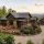 An aerial, wide-angle view of a luxury rustic mountain home with dark wood siding, stone accents, and a metal roof. The property features a spacious wooden deck with outdoor furniture, pristine landscaping, and a concrete driveway leading to a detached two-car garage, set against a backdrop of rolling green mountain ridges at sunset.