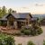 An aerial, wide-angle view of a luxury rustic mountain home with dark wood siding, stone accents, and a metal roof. The property features a spacious wooden deck with outdoor furniture, pristine landscaping, and a concrete driveway leading to a detached two-car garage, set against a backdrop of rolling green mountain ridges at sunset.