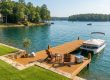 An inviting wooden dock on a calm, sunny lake featuring two wooden Adirondack chairs, a small table with drinks, a cooler, and a pontoon boat moored to the side. The scenic background shows a wide expanse of blue-green water with a few distant boats and a shoreline lined with lush green trees under a clear sky.