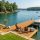 An inviting wooden dock on a calm, sunny lake featuring two wooden Adirondack chairs, a small table with drinks, a cooler, and a pontoon boat moored to the side. The scenic background shows a wide expanse of blue-green water with a few distant boats and a shoreline lined with lush green trees under a clear sky.