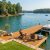 An inviting wooden dock on a calm, sunny lake featuring two wooden Adirondack chairs, a small table with drinks, a cooler, and a pontoon boat moored to the side. The scenic background shows a wide expanse of blue-green water with a few distant boats and a shoreline lined with lush green trees under a clear sky.