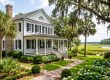 White two-story colonial house with black shutters and a wraparound porch, set in a lush garden near a marsh.