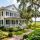 White two-story colonial house with black shutters and a wraparound porch, set in a lush garden near a marsh.