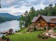 Luxurious lakeside stone house with timber accents, wraparound deck, and a manicured lawn by a calm lake at dusk.