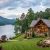 Luxurious lakeside stone house with timber accents, wraparound deck, and a manicured lawn by a calm lake at dusk.
