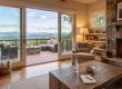 Cozy living room with a stone fireplace, large sliding doors, and a deck overlooking misty blue mountains outdoors. A beige armchair and wooden coffee table sit near a built-in bookshelf and warm lighting.