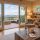 Cozy living room with a stone fireplace, large sliding doors, and a deck overlooking misty blue mountains outdoors. A beige armchair and wooden coffee table sit near a built-in bookshelf and warm lighting.
