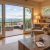 Cozy living room with a stone fireplace, large sliding doors, and a deck overlooking misty blue mountains outdoors. A beige armchair and wooden coffee table sit near a built-in bookshelf and warm lighting.