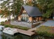 A-frame wooden cabin on a lakeside lot with a large deck, dock, and a boat beside calm water, surrounded by trees.