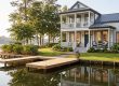 Waterfront two-story home with a wraparound porch, gray siding, and a wooden dock by a calm lake surrounded by trees and landscaping.