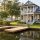 Waterfront two-story home with a wraparound porch, gray siding, and a wooden dock by a calm lake surrounded by trees and landscaping.
