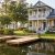 Waterfront two-story home with a wraparound porch, gray siding, and a wooden dock by a calm lake surrounded by trees and landscaping.