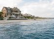 Large coastal house with wraparound porch on a sandy beach, people gathered on the patio.