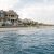 Large coastal house with wraparound porch on a sandy beach, people gathered on the patio.
