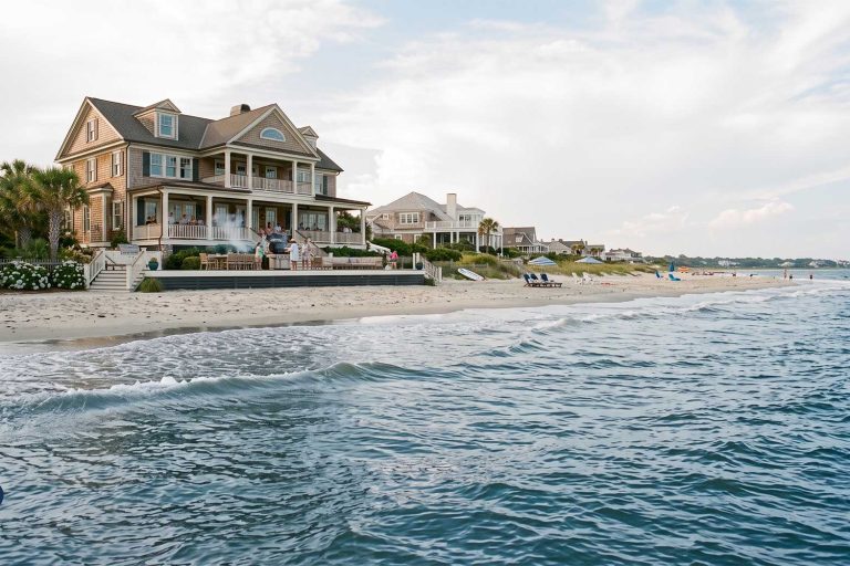Luxury Coastal Home Maintenance for Charleston Getaway Homes Large coastal house with wraparound porch on a sandy beach, people gathered on the patio.