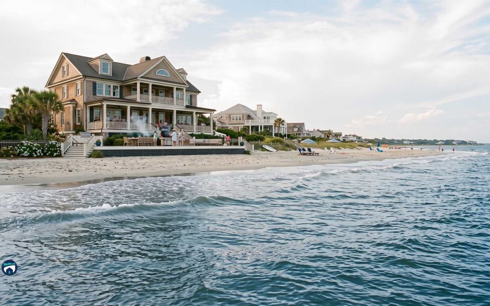 Large coastal house with wraparound porch on a sandy beach, people gathered on the patio.