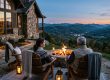Couple sitting in Adirondack chairs around a stone fireplace on a patio, watching a sunset over rolling mountains.