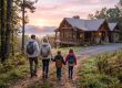 Family of four with backpacks walking toward a wooden cabin on a forest hillside at sunset. (informative)