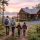 Family of four with backpacks walking toward a wooden cabin on a forest hillside at sunset. (informative)