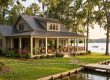 Lakeside cottage with a wraparound porch, hanging plants, and a wooden dock by calm water.