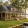 Lakeside cottage with a wraparound porch, hanging plants, and a wooden dock by calm water.