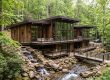 Modern wooden house with expansive glass walls set among dense green forest, built over a rocky, flowing stream.