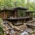 Modern wooden house with expansive glass walls set among dense green forest, built over a rocky, flowing stream.