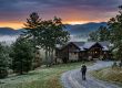 Hiker with backpack walking a gravel path toward a stone-and-wood cabin in a forest at sunset, mountains in the distance.