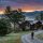 Hiker with backpack walking a gravel path toward a stone-and-wood cabin in a forest at sunset, mountains in the distance.