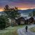 Hiker with backpack walking a gravel path toward a stone-and-wood cabin in a forest at sunset, mountains in the distance.
