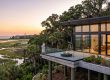 Sunset view from a modern house deck overlooking marshlands; a woman stands by a fire pit near a telescope.