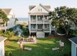 Family and friends gather in a grassy backyard for a beachside barbecue with a grill emitting smoke and a picnic table under a blue sky.