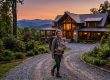 Woman hiking with a baby in a front carrier in front of a wood cabin at sunset on a gravel driveway, mountains in the background.