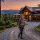 Woman hiking with a baby in a front carrier in front of a wood cabin at sunset on a gravel driveway, mountains in the background.