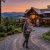 Woman hiking with a baby in a front carrier in front of a wood cabin at sunset on a gravel driveway, mountains in the background.