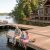 A man and two children fishing on a wooden dock by a lake, with a pontoon boat tied to the dock and a lakeside house in the background. Estate Maintenance Services