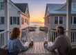 Two people sit at a wooden patio table on a balcony, facing a beach at sunset between two beachfront houses. Estate Manager