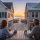 Two people sit at a wooden patio table on a balcony, facing a beach at sunset between two beachfront houses. Estate Manager