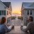Two people sit at a wooden patio table on a balcony, facing a beach at sunset between two beachfront houses. Estate Manager