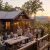 Rustic mountain lodge at sunset with a stone and wood exterior; people sit around a long wooden table on a lit deck, enjoying food and drinks as string lights glow.