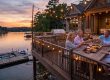 Four adults toast with wine on a lakeside deck at sunset, with string lights and a charcuterie board on the table.