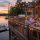 Four adults toast with wine on a lakeside deck at sunset, with string lights and a charcuterie board on the table.