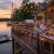 Four adults toast with wine on a lakeside deck at sunset, with string lights and a charcuterie board on the table.
